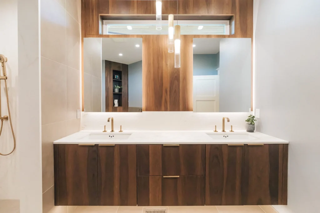 Modern bathroom with dual sinks on a wood vanity, brass fixtures, marble countertop, large mirror, pendant lights, and a small plant on the right side of the counter. Shower controls are visible to the left.