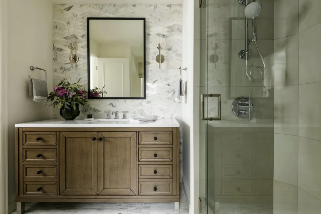 A modern bathroom with a wooden vanity, a white countertop, a vase of flowers, a rectangular mirror, a marble tile backsplash, and a glass-enclosed shower with silver fixtures.