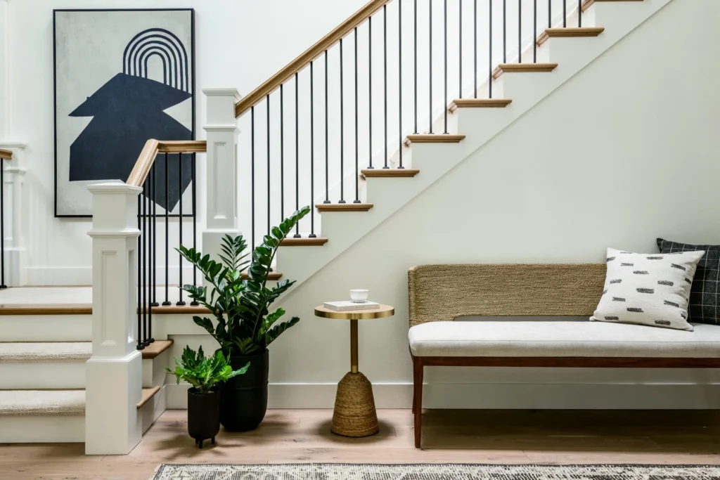 A modern staircase with wooden steps and black railings sits above a small seating area with a beige bench, a pillow, a round side table, two potted plants, and abstract wall art on a white wall.