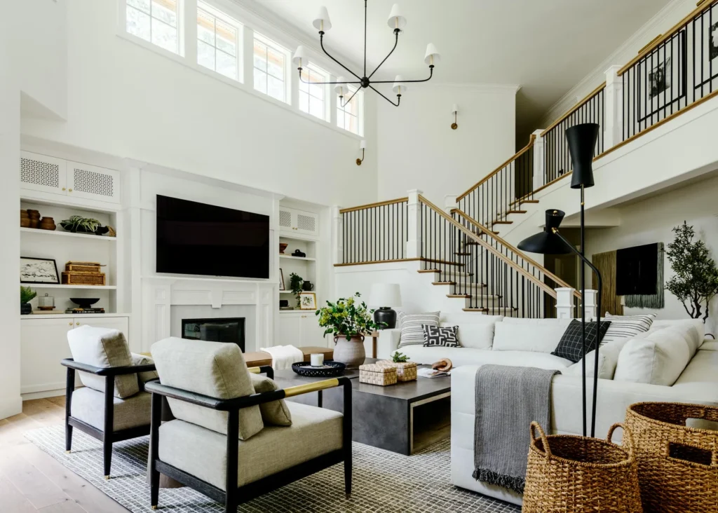 Modern living room with white walls, high ceilings, large windows, a white sectional sofa, two armchairs, a coffee table, and built-in shelves. A staircase with black railings and wooden accents is visible in the background.