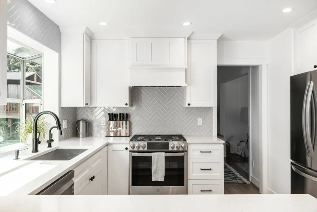 Modern kitchen with white cabinets, stainless steel appliances, a gas stove, herringbone gray backsplash, black faucet, large window, and bright lighting. The counter holds a knife block, kettle, and soap dispenser.
