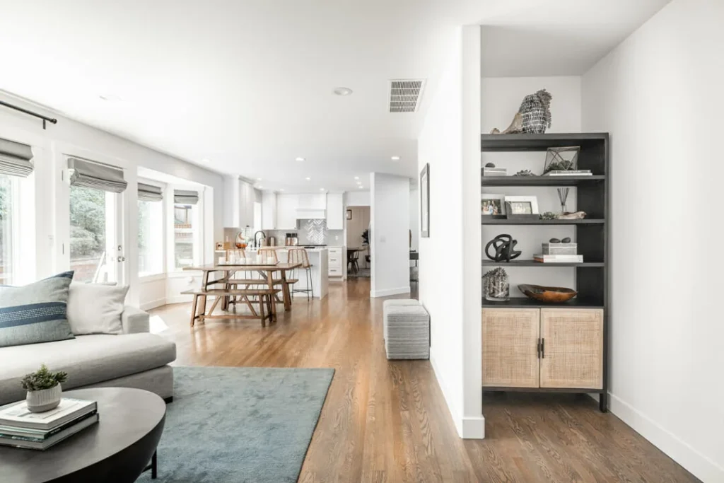 Bright, modern open-concept living and dining area with wooden floors, large windows, cozy gray sofa, dining table, and a black shelving unit with decor items against a white wall.