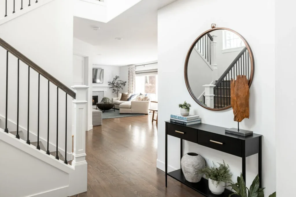 A modern, bright entryway with a round mirror above a black console table holding books, decor, and plants. To the left, a staircase with black railings, and in the background, a cozy living room with a sofa and TV.