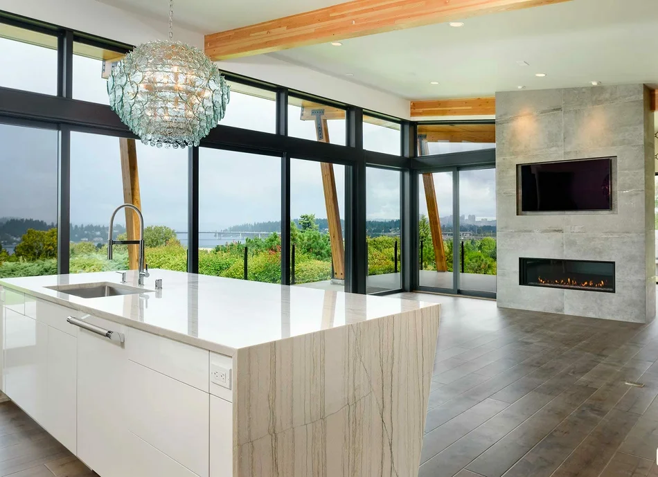 Modern kitchen with a white marble island, large windows offering scenic views, a unique chandelier, built-in fireplace, wall-mounted TV, and wood accents in the ceiling beams.