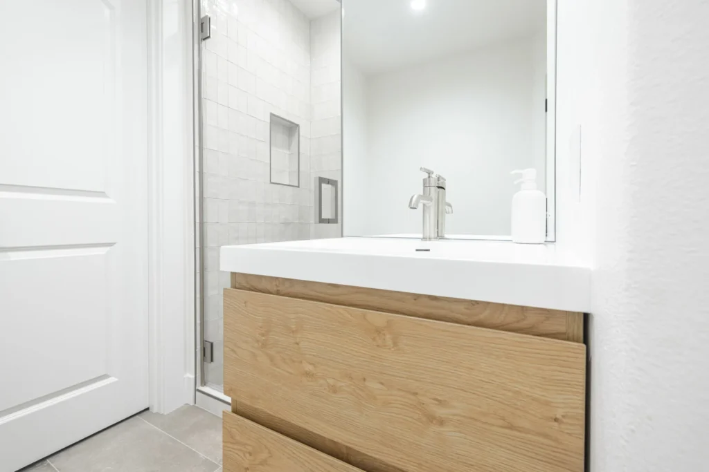 Modern bathroom with a light wood vanity, white countertop, and silver faucet. In the background, there is a glass shower with white tiles and a built-in shelf. A soap dispenser is on the counter.