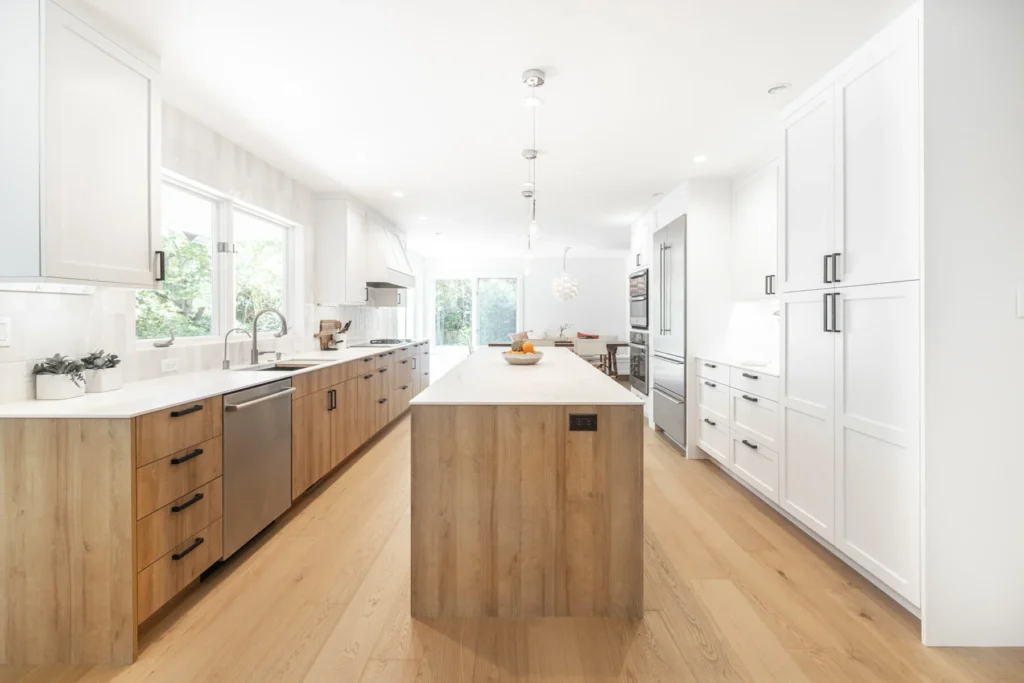 Modern, spacious kitchen with light wood floors, white cabinets, stainless steel appliances, a large island with a sink, and tall windows letting in ample natural light. Fruit bowl and plants decorate the space.