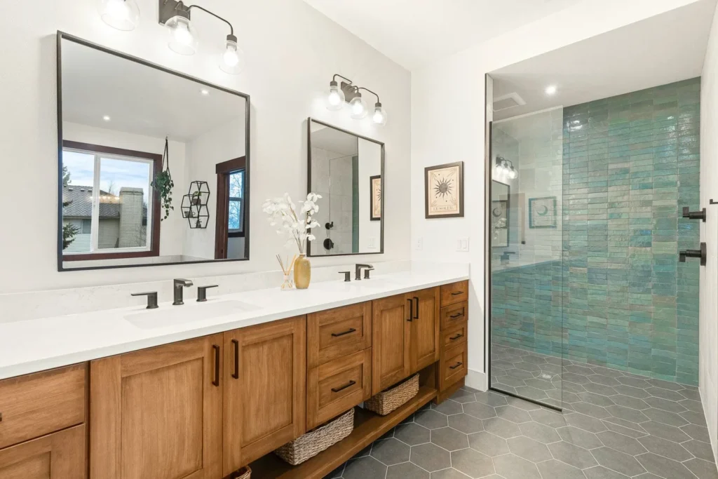 Modern bathroom with a double wood vanity, two mirrors, black fixtures, wall lights, and a large walk-in shower featuring teal tiles and glass door. Gray hexagon floor tiles and decorative accents complete the space.