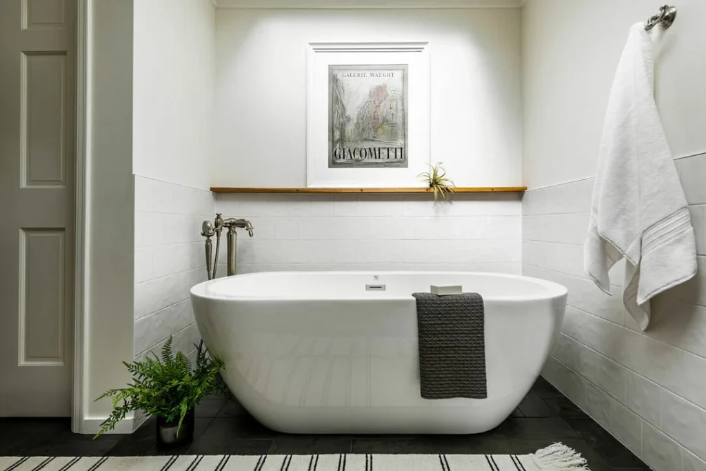 A modern white bathtub with a gray towel draped over the edge sits in a minimalist bathroom. A plant is on the floor, a towel hangs on the wall, and a framed print is displayed above a narrow wooden shelf.